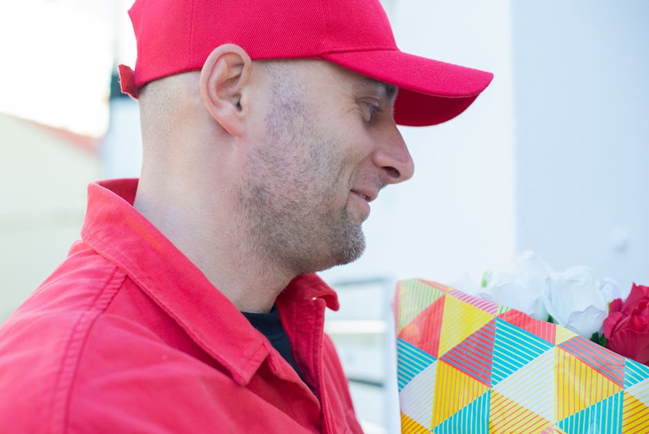 A man wearing a red cap and red jacket is holding a floral gift wrapped in colorful geometric-patterned paper containing white and pink flowers. The bouquet is held close to his face as he looks at it