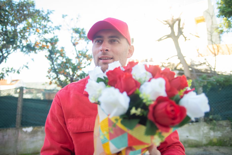 A man dressed in a red courier uniform and matching cap is holding a colorful bouquet of fresh flowers, including red and white roses, wrapped in vibrant paper. He is outdoors, with trees and a fence