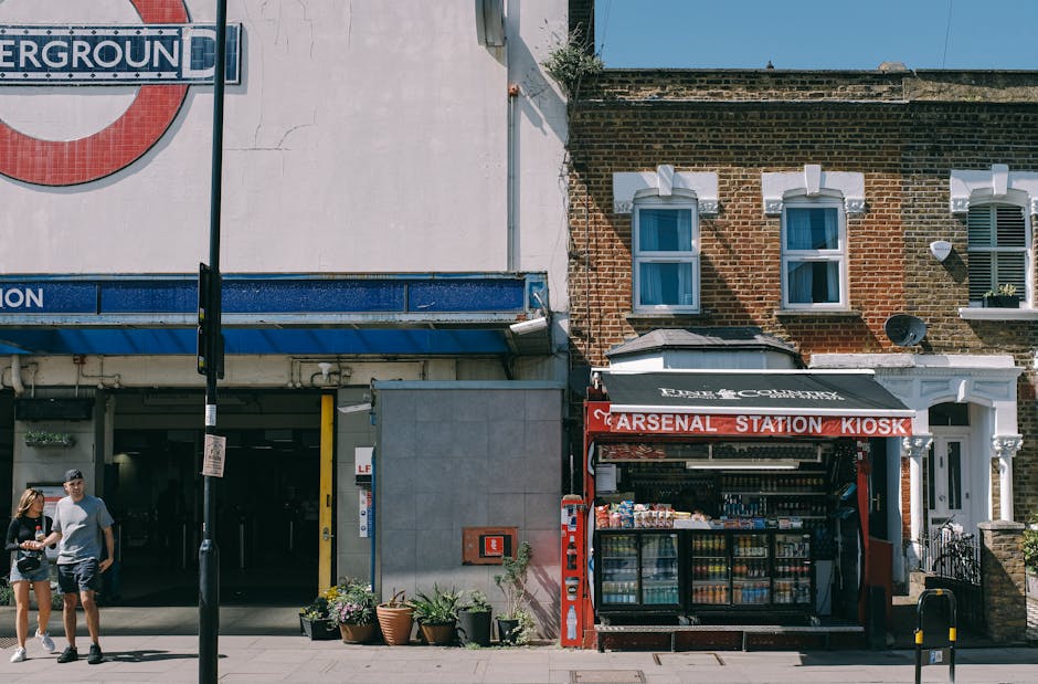 A colorful floral arrangement held by a person in front of the Arsenal Station Kiosk, which is situated on a street with a brick building featuring two white-framed windows and a small awning. The bou