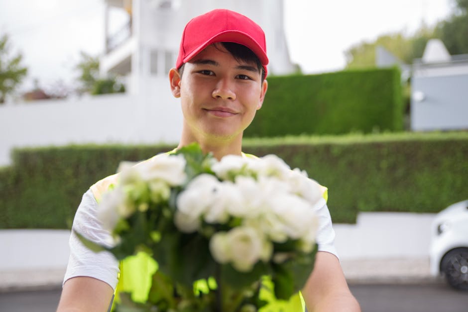A delivery person wearing a red cap and a high-visibility vest is smiling as they hand over a bouquet of fresh white roses and green foliage. The bouquet is arranged in a rounded style, wrapped with l