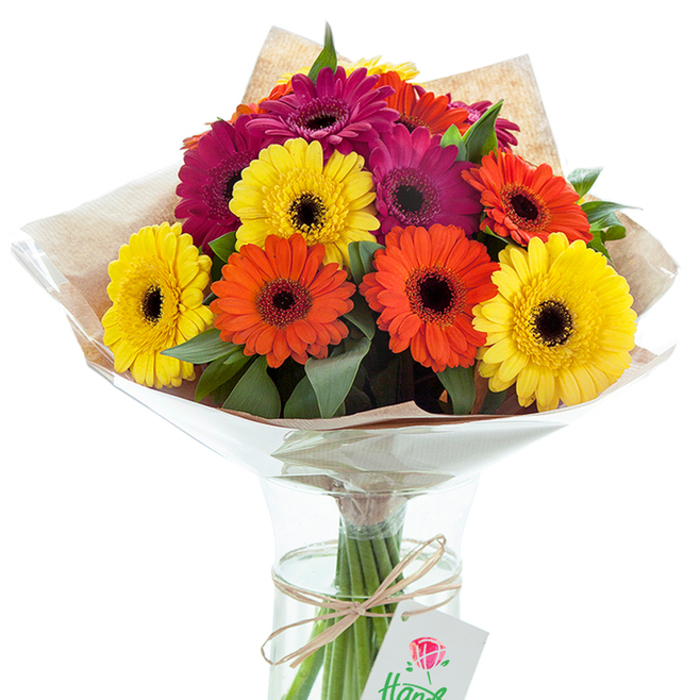 This exquisite hand-tied bouquet, elegantly poised within a transparent glass vase, captures the vibrancy and character of Woolwich with every petal. A constellation of Gerbera daisies-sun-bright yellows, opulent oranges, and rich magenta hues-stands out amid a lush backdrop of verdant green leaves. The unique, dark centers of each bloom provide dramatic contrast and visual rhythm-a touch that mirrors the diversity and energy of the Woolwich community. Encasing the bouquet, a rustic wrap of light brown kraft paper, topped with a layer of crinkled, protective cellophane, enhances both structure and style, as if echoing the tactile charm of Woolwich's weekend market stalls. The vase reveals an artful tangle of green stems in pure water, reinforcing the bouquet's freshness and vibrancy. A simple bow of country-style twine is tied around the neck, from which a small, crisp white tag with a delicate red rose drawing hangs, signifying a locally crafted gift. Subtle daylight bathes the entire composition, amplifying the radiant hues and highlighting the impeccable arrangement. This image, with its celebratory spirit and expert composition, is ideal for illustrating stories about local gift-giving, flower delivery services, or special occasions cherished near Woolwich Dockyard or along the Thames Path. It's a beautiful reminder of the joy that thoughtfully arranged flowers can bring to any home or event in Woolwich.